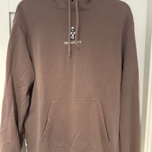 Brun hoodie med tryck - Säljer en brun hoodie med ett litet tryck på bröstet. Den har en klassisk känguruficka och justerbar huva med snören. Perfekt för en avslappnad stil. Den är i storlek M