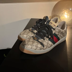 Gucci sneakers med monogram - Snygga Gucci sneakers med det klassiska monogrammönstret. Skorna har gröna och röda ränder på sidan och svarta snören. Perfekta för en stilren look.