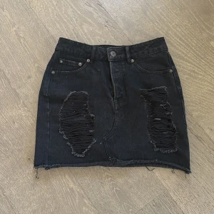 Svart lågmidjad jeanskjol med slitningar - Snygg svart lågmidjad jeanskjol med slitna detaljer framtill. Kjolen har en klassisk femficksdesign och en rå kant nedtill för en edgy look. Perfekt för en avslappnad stil.