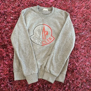 Monclear tröja - Säljer en stilren grå sweatshirt från Moncler med ett stort, iögonfallande logotryck i rött och grått på framsidan. Bara använd några gånger. Storlek M. Är äkta köpt för ett år sedan men inte mycket använd.