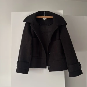 Mörkgrå kappa med stora fickor - Snygg mörkgrå kappa med stora fickor och knappar framtill. Den har en klassisk krage och långa ärmar med uppvikta muddar. Perfekt för kyligare dagar. Från kappahl i storlek S