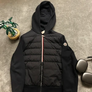 Svart jacka med huva från Moncler - Säljer en stilren svart jacka från Moncler med huva och dragkedja. Jackan har en quiltad framsida och Moncler-logga på ärmen. Perfekt för kyligare dagar.