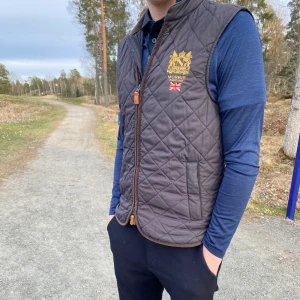Brun quiltad väst från Morris - Säljer en snygg brun quiltad väst från Morris med dragkedja och broderad logga på bröstet. Perfekt för en stilren look. Västen har en klassisk design med en liten krage och två fickor framtill.