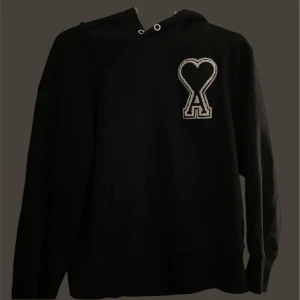 AMI - Snygg svart hoodie med en unik hjärtdetalj på bröstet. Perfekt för en avslappnad stil med en touch av personlighet. Den har långa ärmar och en klassisk huva med dragsko.