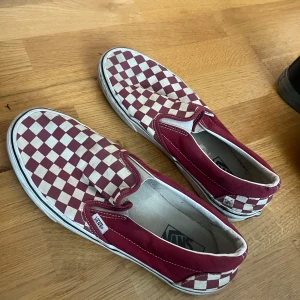 Röd och vit rutiga slip-on sneakers från Vans - Säljer ett par klassiska slip-on sneakers från Vans i röd och vit rutig design. Skorna har en bekväm passform och är perfekta för en avslappnad stil. De är lätta att ta på och av tack vare den elastiska sidan.