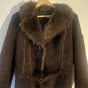 Brun mockajacka med pälsdetaljer från Zara - Säljer en snygg brun mockajacka från Zara med mjuka pälsdetaljer runt kragen. Jackan har knappar och ett bälte i midjan för en justerbar passform. Perfekt för kyligare dagar. Köpt secondhand och den är sååå fin men kommer inte till användning