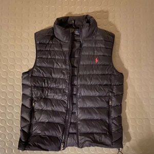  Ralph Lauren Väst - Snygg svart dunväst från Ralph Lauren med dragkedja och broderad logga i rött på bröstet. Perfekt för lager-på-lager under kyliga dagar. Västen har en quiltad design och två praktiska fickor med dragkedja. Hör av er vid frågor