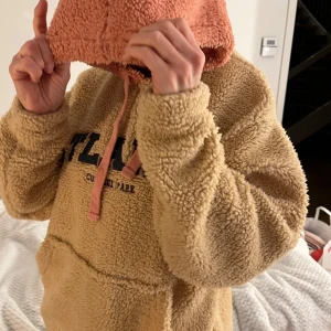Beige teddy-hoodie från ASOS DESIGN - Mysig beige teddy-hoodie från ASOS DESIGN med texten 'ATLANTA' på bröstet. Hoodien har en kontrasterande rosa huva och en stor magficka. Perfekt för kyliga dagar.