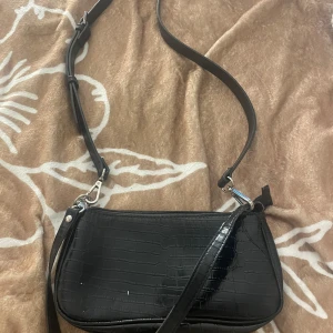 Svart axelväska - Snygg svart axelväska med krokodilpräglat mönster. Väskan har en justerbar axelrem och stängs med dragkedja. Minns inte nypriset på väskan