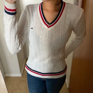 Vit stickad tröja från Tommy Hilfiger - Säljer en vit stickad tröja från Tommy Hilfiger med v-ringning och kabelstickat mönster. Tröjan har långa ärmar och ribbade muddar med röd och marinblå randdetalj vid ärmslut och nederkant. Perfekt för en stilren look!