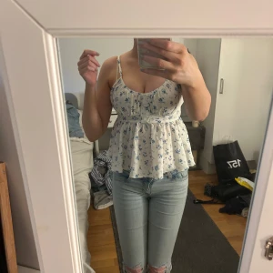 Blommig vit linnetopp - Säljer en söt vit linnetopp med blå blommor. Toppen har smala axelband och en knytning i ryggen. Perfekt för sommardagar!