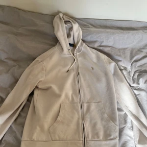 Beige hoodie från Ralph Lauren - Säljer en stilren beige hoodie från Ralph Lauren med dragkedja och broderad logga på bröstet. Tröjan har långa ärmar och en praktisk huva med snörning. Köpte för en månad sen för 1600kr och knappt använd. Priset är inte hugget i sten och bara att höra av sig vid frågor 