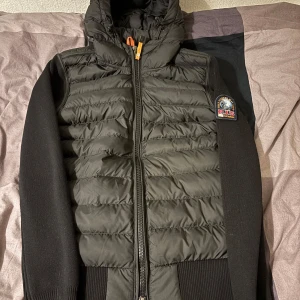 Svart pufferjacka från Parajumpers - Säljer en svart pufferjacka från Parajumpers med huva och dragkedja. Jackan har ett emblem på ärmen och ribbade muddar. Perfekt för kyliga dagar.