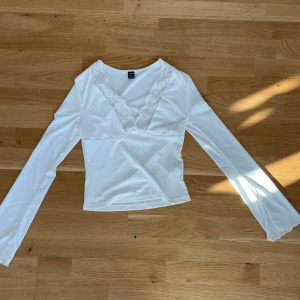 Vit långärmad topp med spetsdetaljer från Shein - Säljer en stilren vit topp från Shein med vackra spetsdetaljer vid den v-ringade halsen. Toppen har långa ärmar och en mjuk, bekväm passform. Perfekt för att ge en elegant touch till din outfit.