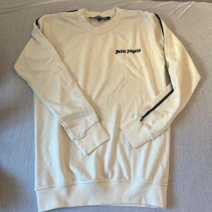 Vit långärmad tröja från Palm Angels - Säljer en stilren vit långärmad tröja från Palm Angels med svart logotyp på bröstet och svarta ränder längs ärmarna. Perfekt för en avslappnad och trendig look.