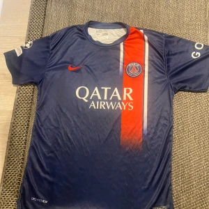  PSG fotbollströja - Säljer en blå PSG fotbollströja från Nike med korta ärmar. Tröjan har röda och vita detaljer samt sponsorloggor. Perfekt för fans av laget!
