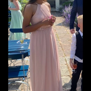 Ljusrosa långklänning - Säljer min fina ljusrosa långklänning med halterneck och vackra veckdetaljer. Klänningen har en smickrande passform med ett band i midjan dekorerat med pärlor. I ryggen är även lite öppen. Perfekt klänning för sommar event eller bal🥰