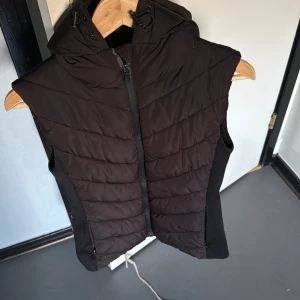 Svart dunväst från Zara - Snygg svart dunväst från Zara med dragkedja och huva. Perfekt för kyliga dagar när du vill hålla värmen utan att kompromissa med stilen. Västen har en quiltad design som ger ett modernt och sportigt utseende.