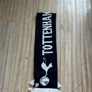 Tottenham halsduk  - Snygg halsduk i mörkblått med vit text och logga för Tottenham. Perfekt för att visa ditt stöd för laget. Halsduken har fransar i ändarna och är gjord i ett mjukt material. Köpt på arenan Tottenham hotspurs ny pris 200kr säljer för 65kr