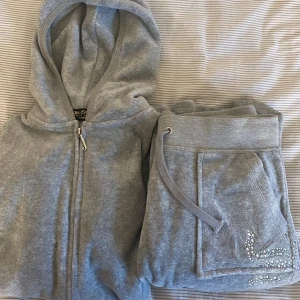 Grå hoodie och byxor från Juicy Couture - Säljer en grå hoodie och matchande byxor från Juicy Couture. Hoodien har dragkedja och en luva, medan byxorna har en praktisk ficka och dragsko i midjan. Båda plaggen är dekorerade med glittriga detaljer och texten 'Juicy Couture' på baksidan av hoodien.
