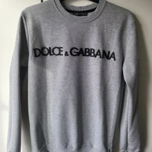 Dolce & gabbana tröja  - Säljer en stilren grå sweatshirt från Dolce & Gabbana med deras logga i svart på bröstet. Tröjan har långa ärmar och en rund halsringning med ribbade kanter. Perfekt för en avslappnad och trendig look.
