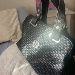Svart vintage Valentino handväska  - Snygg svart handväska med silverdetaljer och dragkedja. Perfekt för att bära dina dagliga nödvändigheter med stil. Väskan har två handtag och en rymlig insida.