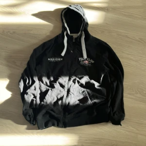 Svart hoodie från polo club - Säljer en svart hoodie från Saint Vincent med dragkedja och tryck på bröstet. Hoodien har vita snören och en klassisk design med texten 'Polo Club' och 'Saint Vincent' på framsidan. Perfekt för en avslappnad stil.