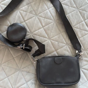 Svart axelväska med rund plånbok - Snygg svart axelväska i syntetmaterial med en matchande rund plånbok. Väskan har en justerbar axelrem och stängs med dragkedja. Perfekt för att hålla dina saker organiserade och stilrena.