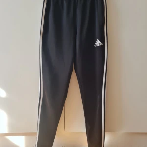 Svarta Adidasbyxor - Snygga svarta träningsbyxor från Adidas med 3 vita ränder längs sidorna. Byxorna har en elastisk midja för extra komfort och är perfekta för träning eller en avslappnad dag. Storlek S. Kan hämtas i Vallentuna.