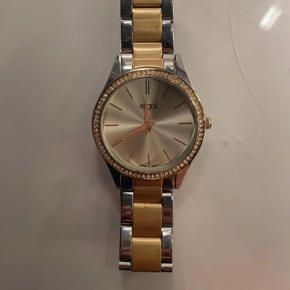 Elegant klocka från Regal med en kombination av guld och silver i armbandet. Urtavlan är omgiven av en glittrig kant och har en stilren design med guldiga visare och markeringar. Perfekt accessoar för att lyfta din stil. (pris kan diskuteras) . Asusteet.