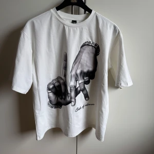 Vit t-shirt med tryck - Riktigt snygg vit t-shirt med ett coolt svartvitt tryck av händer med ringar. Perfekt för en avslappnad stil. T-shirten är oversized. Storlek: S. Vet ej vilket märke det är då jag köpte den second hand helt ny. Jag har själv aldrig använt den och alltså är den helt oanvänd. 