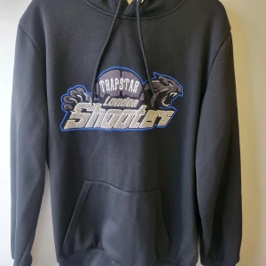 Trapstar hoodie - Säljer en svart hoodie från Trapstar med tryck på framsidan. Hoodien har en stor ficka framtill och justerbar huva med dragsko. Perfekt för en casual look.