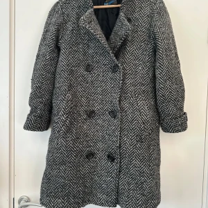 Grå och svart kappa med fiskbensmönster - Snygg grå och svart kappa med fiskbensmönster och dubbelknäppning. Den har långa ärmar och stora knappar framtill. Perfekt för vår/höst. 