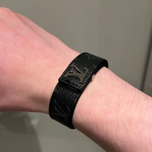 Svart Louis Vuitton armband  - Snyggt svart armband i skinn från Louis Vuitton med det ikoniska LV-märket i silver. Armbandet har ett stilrent mönster och justerbar passform. Perfekt för att ge din outfit en lyxig touch.