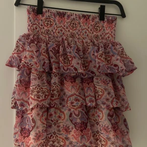 Mönstrad kjol från Lindex - Säljer en färgglad kjol från Lindex med ett vackert mönster i rosa, lila och rött. Kjolen har volanger och en elastisk midja för en bekväm passform. Perfekt för att liva upp garderoben!