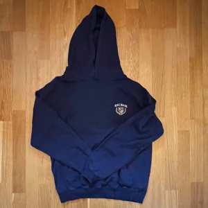 Mörkblå hoodie från Urban Outfitters  - Säljer en mörkblå hoodie från Urban Outfitters med en liten logga på bröstet. Tröjan har en klassisk design med huva och långa ärmar. 
