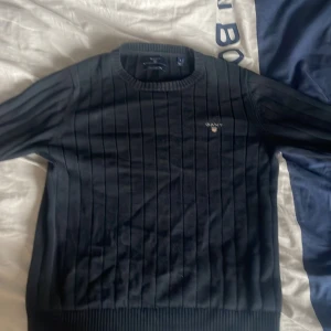 Svart ribbad tröja från GANT - Säljer en stilren svart tröja från GANT i premium bomull. Tröjan har ett ribbat mönster och GANT-loggan broderad på bröstet. Perfekt för en klassisk och tidlös look.