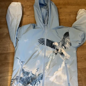 Ljusblå jacka med örntryck från Trapstar - Säljer en cool ljusblå jacka från Trapstar med ett stort örntryck och text på ryggen. Jackan har dragkedja och huva, perfekt för en streetwear-look. Den har långa ärmar och två fickor med dragkedja framtill. Storlek S, om ni undrar något hör av er!