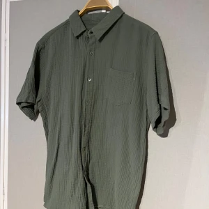 Grön kortärmad skjorta från S.C.W. - Snygg overshirt och härlig grön färg. Färgen är en aning mörkare i verkligheten! Materialet är bomull men det påminner mer om linnetyg, så perfekt till sommaren.