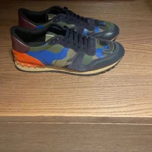 Valentino Rockrunner  - Snygga Valentino Rockrunner sneakers i ett coolt camouflage-mönster med inslag av blått och orange. Skorna har en klassisk snörning och en robust sula för extra komfort. Perfekta för dig som vill sticka ut med stil.