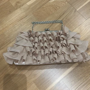 Beige fransväska från Tiamo - Beige fransväska från Tiamo med volangdetaljer och en kedjehandtag. Väskan har en dragkedja inuti för förvaring.🩷🩷