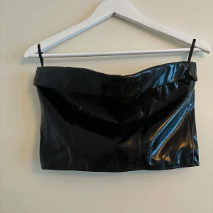 Svart glansig tubtopp med dragkedja - Snygg helt oanvänd svart glansig tubtopp med dragkedja framtill från GinaTricot. Perfekt för en utekväll. Den har en stilren design med en bred kant upptill. Ursprungspris 399kr