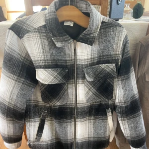 Rutig jacka från Zara - Säljer en snygg rutig jacka från Zara i svart, grått och vitt. Jackan har en dragkedja framtill och två bröstfickor med lock. Perfekt för höst och vår!