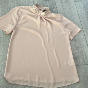 Beige blus med pärldetaljer - Elegant beige blus  med korta ärmar och en hög krage dekorerad med pärlor i guld och vitt. Perfekt för en stilren look.