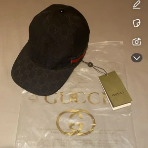 Gucci keps - Snygg svart keps från Gucci med klassiskt GG-monogram över hela kepsen och en ikonisk grön-röd rand på sidan. Kepsen har böjd skärm och justerbar passform. Perfekt för dig som vill ha en exklusiv och trendig accessoar.
