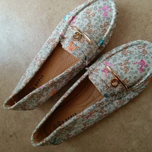 Färgglada loafers med gulddetalj - Snygga loafers från Dapple i beige tyg med färgglada bokstäver och guldfärgad metallspänne framtill. Skorna har platt sula med blommönster undertill och rund tå. Perfekta för dig som gillar unika detaljer och bekväm stil.