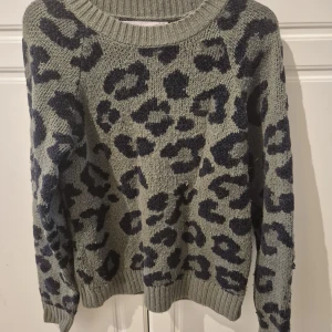 Leopardmönstrad stickad tröja ONLY - Grå stickad tröja från ONLY med mörkblått leopardmönster. Tröjan har rund halsringning, ribbade muddar och lång ärm. Perfekt för dig som vill sticka ut med en trendig och cool look.