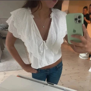 Vit blus med broderade volangärmar - Supersöt vit blus med djup v-ringning, knyt framtill och breda volangärmar med broderade detaljer. Luftig och croppad modell som ger en romantisk vibe. Perfekt till jeans eller kjol för en trendig look.