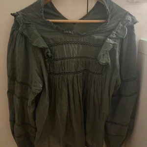 Olivgrön blus med volangdetaljer - Säljer en väldigt fin olivgrön blus med volangdetaljer vid axlarna och broderade sömmar. Blusen har en lös passform, långärmade ärmar och är tillverkad i ett lätt, vävt material som är mycket skönt. Perfekt för dig som gillar boho chic-vibe och snygga detaljer!!🥰