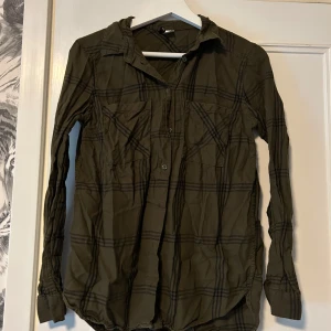 Olivgrön rutig skjorta  - Olivgrön skjorta med svart rutigt mönster, klassisk krage och två bröstfickor. Långärmad modell med knappar framtill och vid ärmslut. Skjortan har en avslappnad passform och är tillverkad i mjukt bomullstyg. Perfekt för en chill och stilren look.